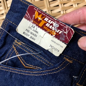 May include: A close-up of a dark blue denim jean pocket with a brown label that reads "RAPID TRANSIT Wrangler" and "28 L (71) LONG PRE-WASH BOOT JEAN" and "100% COTTON 7M542PW".