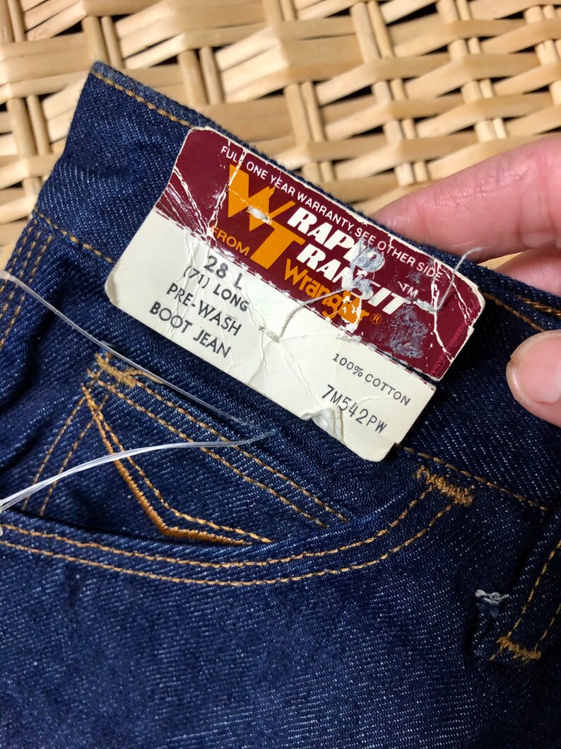 May include: A close-up of a dark blue denim jean pocket with a brown label that reads "RAPID TRANSIT Wrangler" and "28 L (71) LONG PRE-WASH BOOT JEAN" and "100% COTTON 7M542PW".