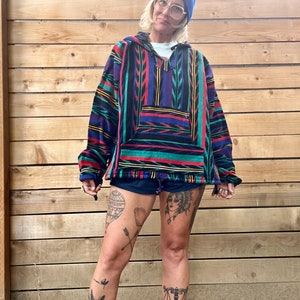 May include: A person wearing a blue beanie, glasses, and a colourful striped fleece hoodie with a pocket. The hoodie has a geometric pattern with red, blue, green, and yellow stripes. The person is wearing black shorts and has several tattoos on their legs.