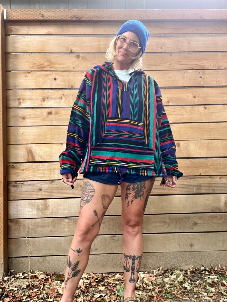 May include: A person wearing a blue beanie, glasses, and a colourful striped fleece hoodie with a pocket. The hoodie has a geometric pattern with red, blue, green, and yellow stripes. The person is wearing black shorts and has several tattoos on their legs.