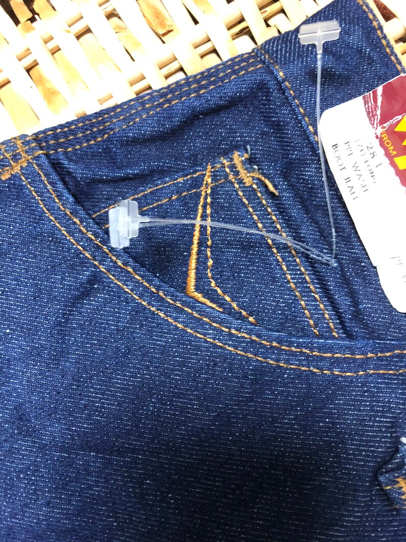 May include: A pair of dark blue denim jeans with a brown stitch detail. The jeans have a small pocket on the right side and a tag attached with the text "28" "1" "IN WAIST" "BOOT CUT" "TALL".