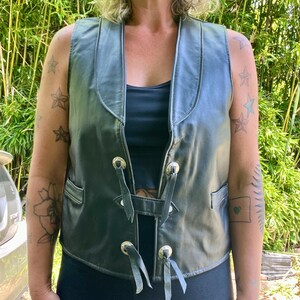 May include: Black leather vest with a V-neck and decorative silver-toned metal accents. The vest has a lace-up closure and small pockets. The sleeveless design is suitable for layering. The vest is worn over a black top.