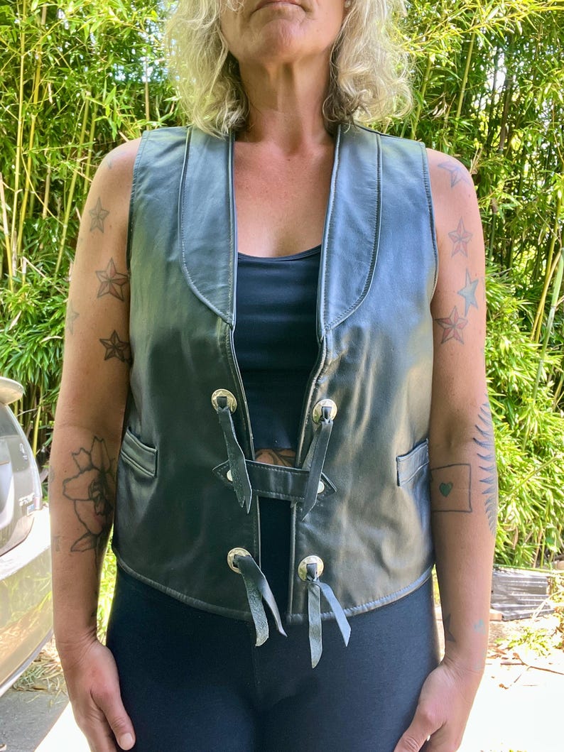 May include: Black leather vest with a V-neck and decorative silver-toned metal accents. The vest has a lace-up closure and small pockets. The sleeveless design is suitable for layering. The vest is worn over a black top.