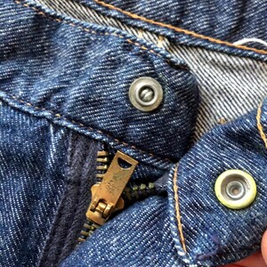 May include: Close-up of a blue denim zipper fly with a metal zipper pull and two metal buttons. The zipper is partially open and the buttons are visible.