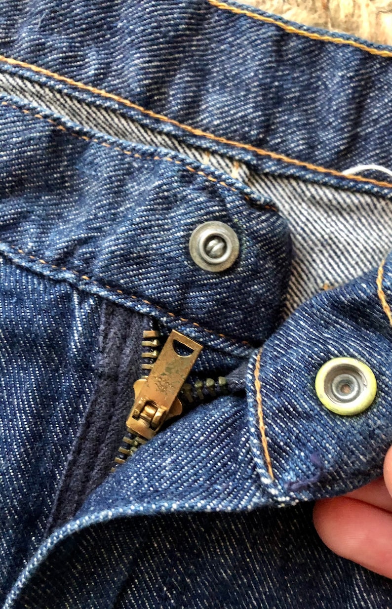 May include: Close-up of a blue denim zipper fly with a metal zipper pull and two metal buttons. The zipper is partially open and the buttons are visible.