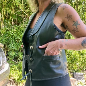 May include: Black leather vest with a notched collar, decorative metal buttons, and front pockets. The vest has a lace-up detail on the sides and is worn over a black top. The vest is a classic piece of outerwear.