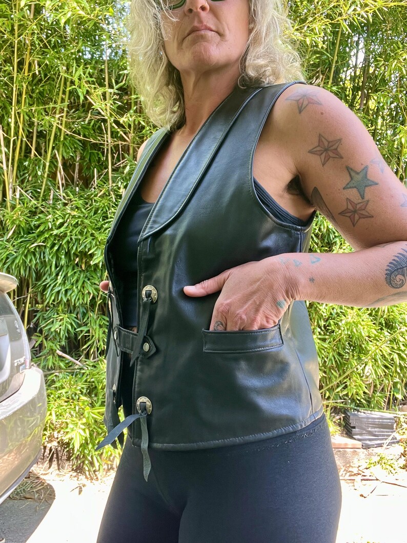 May include: Black leather vest with a notched collar, decorative metal buttons, and front pockets. The vest has a lace-up detail on the sides and is worn over a black top. The vest is a classic piece of outerwear.