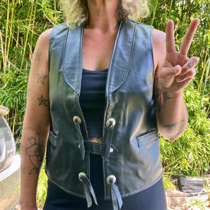 May include: Black leather vest with silver-toned hardware and black ties. The vest has a V-neck and two front pockets. The person in the image is wearing a black top and black leggings. The background is a green bamboo plant.