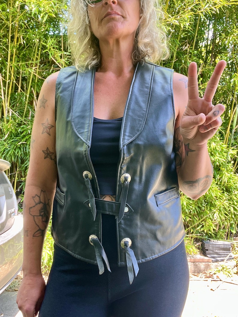 May include: Black leather vest with silver-toned hardware and black ties. The vest has a V-neck and two front pockets. The person in the image is wearing a black top and black leggings. The background is a green bamboo plant.