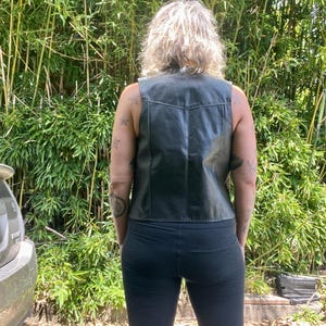 May include: Black leather vest with a classic design. The vest has a V-neck and a tailored fit. The person is wearing black leggings. The background features lush green bamboo.