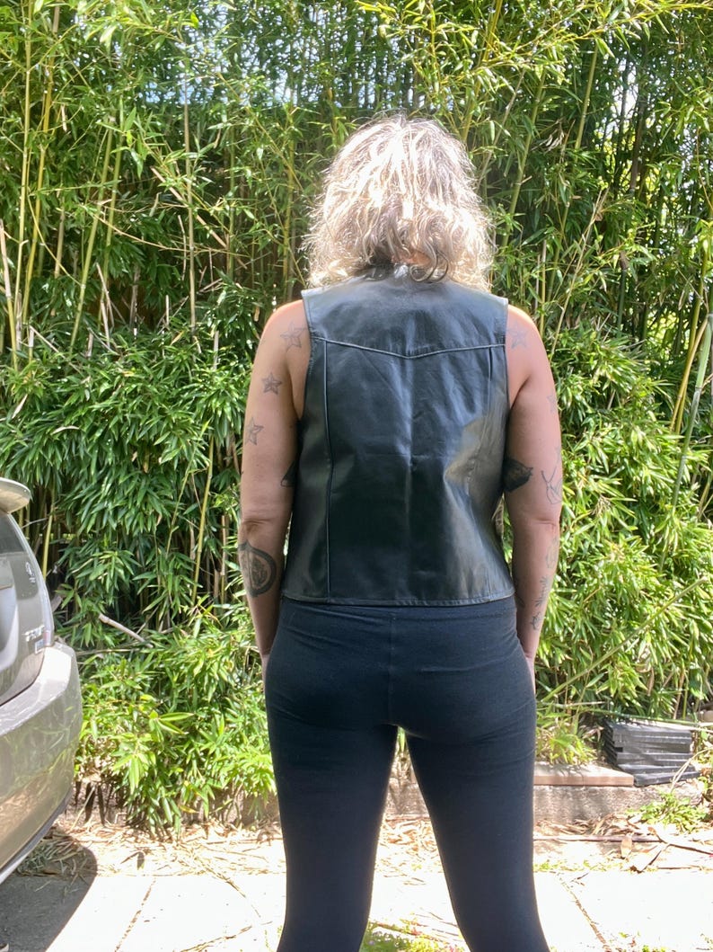 May include: Black leather vest with a classic design. The vest has a V-neck and a tailored fit. The person is wearing black leggings. The background features lush green bamboo.