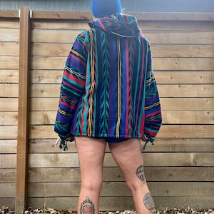 May include: A person wearing a blue beanie and a colourful striped hooded jacket. The jacket has a geometric pattern with red, yellow, blue, and green stripes. The person is wearing shorts and has tattoos on their legs.
