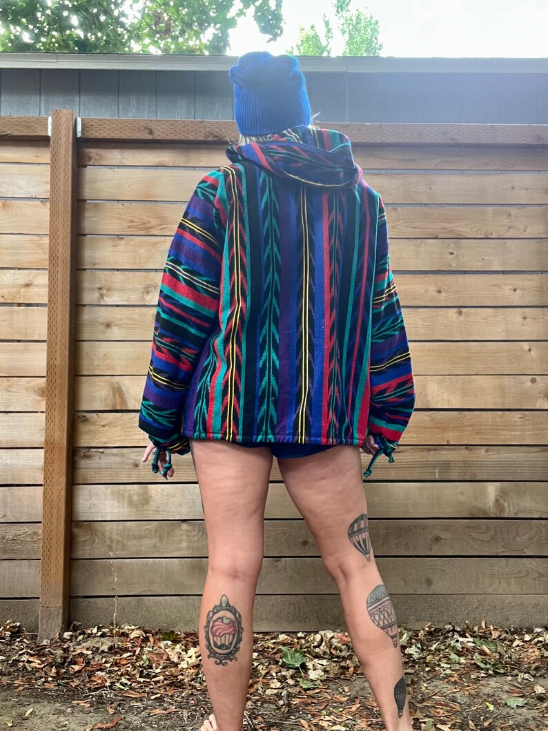 May include: A person wearing a blue beanie and a colourful striped hooded jacket. The jacket has a geometric pattern with red, yellow, blue, and green stripes. The person is wearing shorts and has tattoos on their legs.