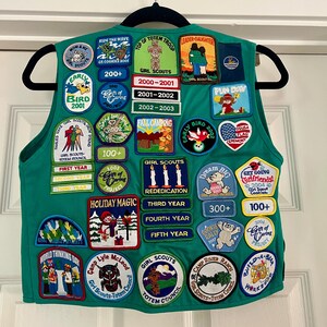 May include: A green Girl Scout vest adorned with numerous colorful patches. Patches feature text like "Girl Scouts," "Holiday Magic," and "Early Bird." Patches also display images of a totem, a bear, and a flag. The vest is hanging on a black hanger.