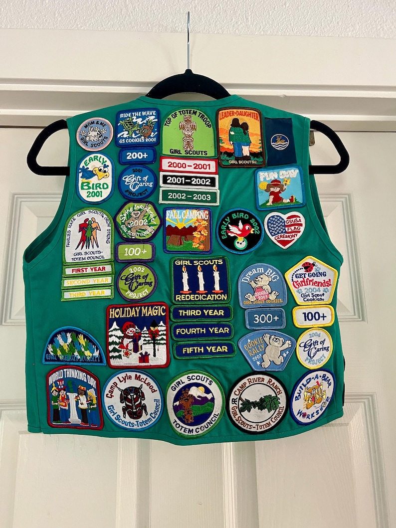 May include: A green Girl Scout vest adorned with numerous colorful patches. Patches feature text like "Girl Scouts," "Holiday Magic," and "Early Bird." Patches also display images of a totem, a bear, and a flag. The vest is hanging on a black hanger.