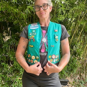 May include: A teal Girl Scout vest adorned with various patches and pins. The vest is worn over a dark gray t-shirt with a colorful graphic and the text "NOTHING PHASES ME". The person is wearing black shorts and glasses.