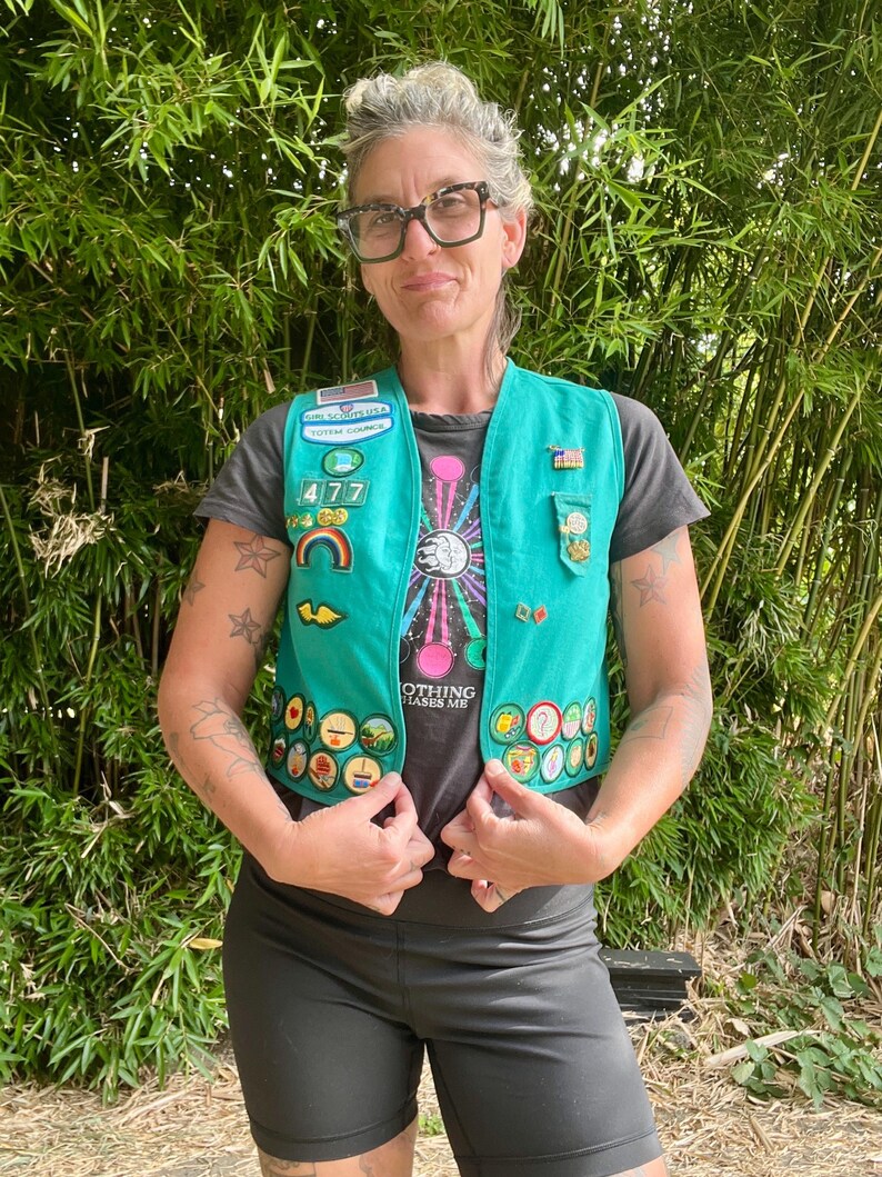 May include: A teal Girl Scout vest adorned with various patches and pins. The vest is worn over a dark gray t-shirt with a colorful graphic and the text "NOTHING PHASES ME". The person is wearing black shorts and glasses.