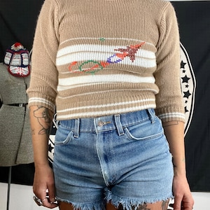 May include: A brown sweater with white stripes and a colorful geometric design. The sweater has a crew neck and 3/4 sleeves.