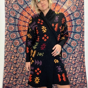 May include: Black knitted cardigan with a geometric pattern in shades of orange, yellow, purple, and green. The cardigan has a shawl collar and a long, loose fit.