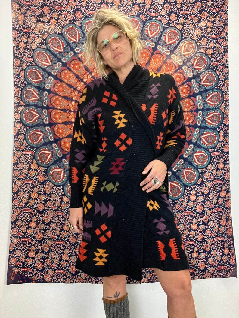 May include: Black knitted cardigan with a geometric pattern in shades of orange, yellow, purple, and green. The cardigan has a shawl collar and a long, loose fit.