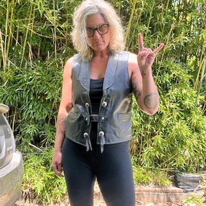 May include: A black leather vest with silver-colored buttons and a buckle closure. The vest has a V-neck and small pockets. The person is wearing black leggings and a black top. The background is a lush green bamboo forest.
