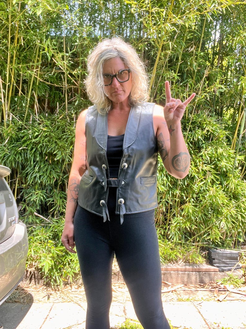 May include: A black leather vest with silver-colored buttons and a buckle closure. The vest has a V-neck and small pockets. The person is wearing black leggings and a black top. The background is a lush green bamboo forest.