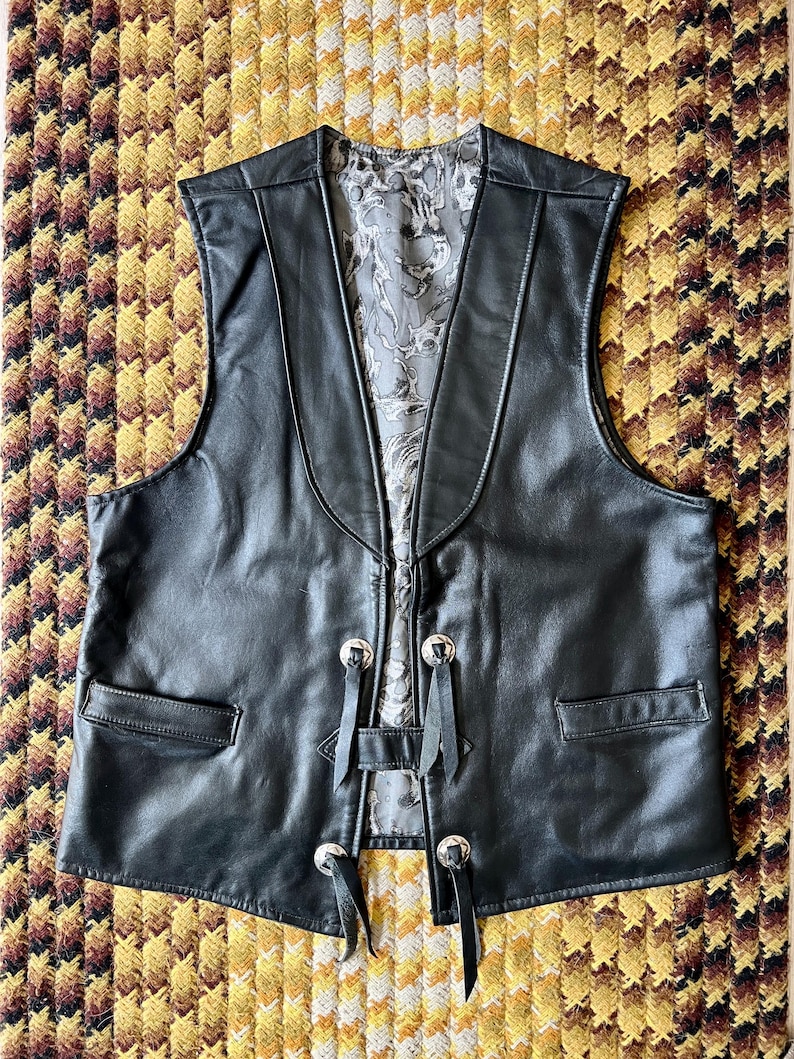 May include: A black leather vest with a notched lapel and two front pockets. The vest has silver-toned buttons and decorative black leather ties. The interior lining features a gray and white floral pattern. The vest is displayed on a patterned yellow and brown surface.