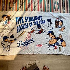 Peut inclure: Serviette de plage blanche avec un logo bleu et rouge des Los Angeles Dodgers et le texte "Five Straight Rookies of the Year". La serviette représente cinq joueurs de baseball portant des uniformes des Dodgers.