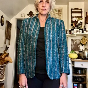 May include: A teal blue blazer with a striped pattern. The blazer has a mandarin collar and long sleeves. The person wearing the blazer is standing in a kitchen.