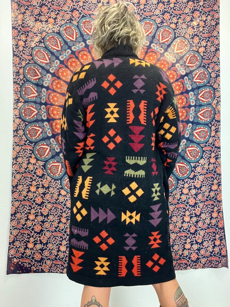 May include: A long black cardigan with a geometric pattern in shades of orange, yellow, red, purple, and green. The pattern is reminiscent of traditional Native American designs.