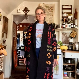 May include: A woman wearing a black cardigan with a colorful geometric pattern. The cardigan has a shawl collar and long sleeves. The woman is also wearing blue jeans and a light blue t-shirt with the text "Doodger" and "my fam".