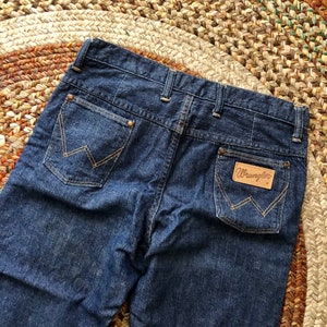 May include: A pair of dark blue denim jeans with a brown leather Wrangler label on the back pocket.