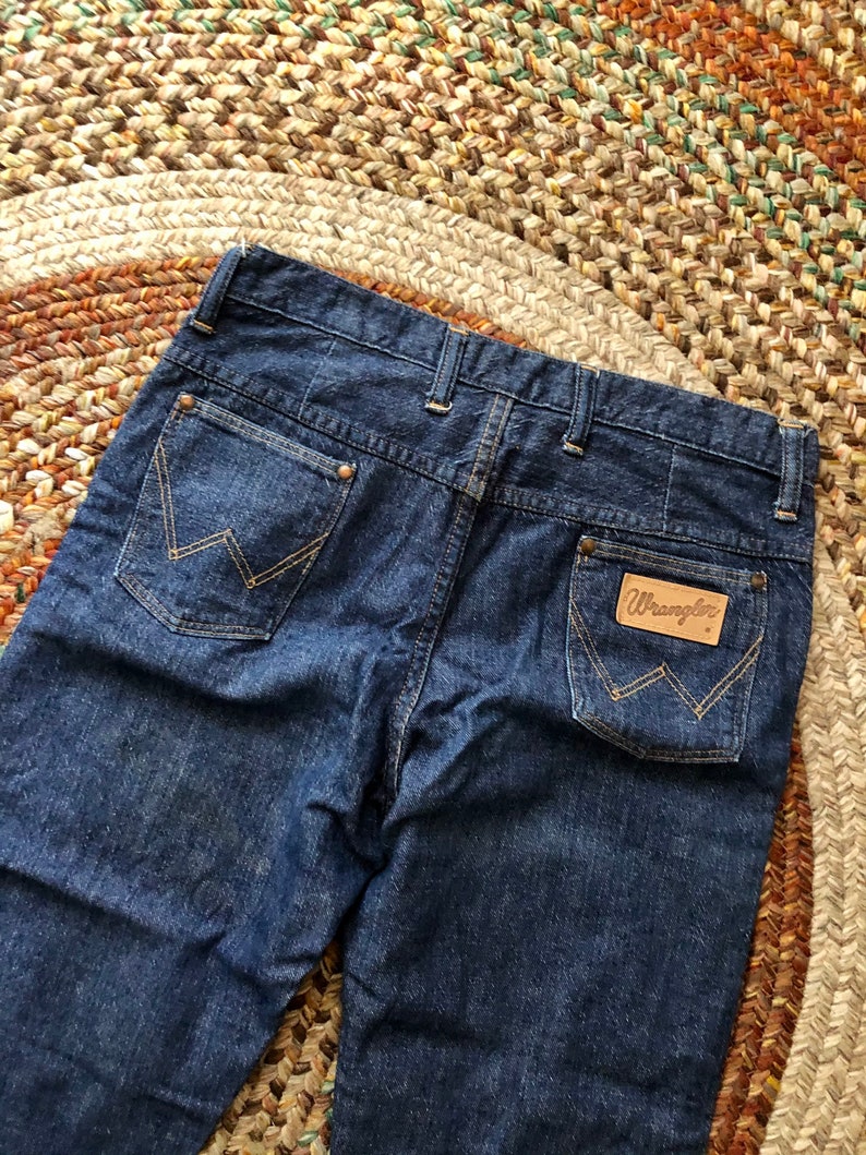 May include: A pair of dark blue denim jeans with a brown leather Wrangler label on the back pocket.