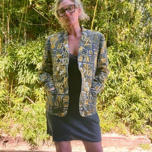 May include: A woman wearing a green and gold patterned blazer with a black sleeveless top underneath. She is standing in front of a green leafy background.
