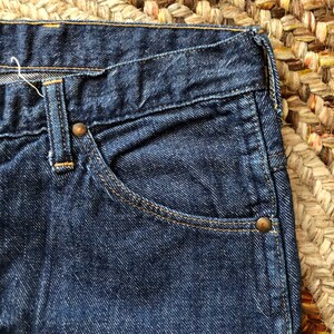 May include: Close-up of a dark blue denim pocket with a copper rivet. The pocket is part of a pair of jeans.