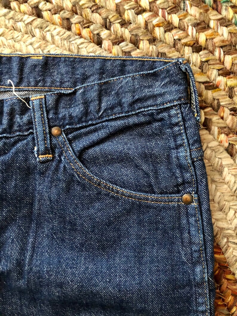 May include: Close-up of a dark blue denim pocket with a copper rivet. The pocket is part of a pair of jeans.