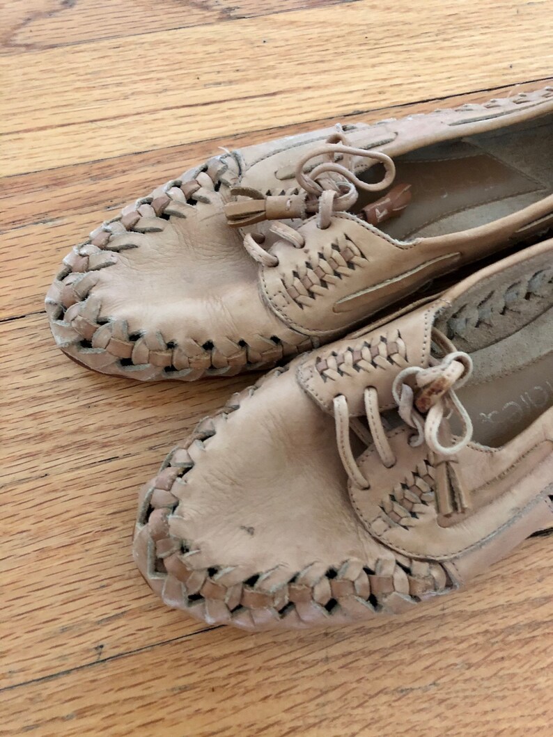 May include: A pair of tan leather shoes with woven detailing and laces. The shoes have a tassel accent on the laces.