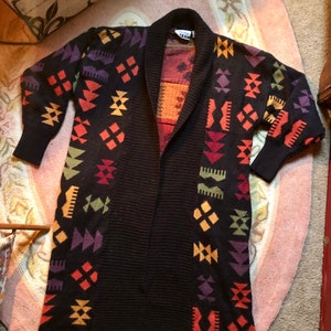 May include: A long, black cardigan with a geometric pattern in shades of orange, yellow, green, purple, and red. The cardigan has a shawl collar and is open in the front.
