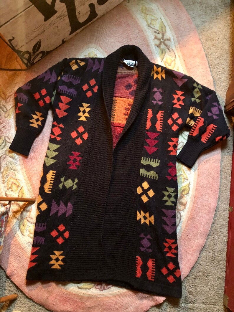May include: A long, black cardigan with a geometric pattern in shades of orange, yellow, green, purple, and red. The cardigan has a shawl collar and is open in the front.