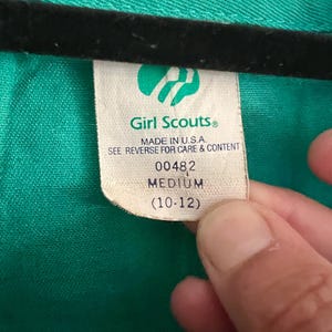 May include: A close-up of a green garment with a tag. The tag reads "Girl Scouts&reg; MADE IN U.S.A." and indicates a medium size (10-12). The garment is a solid green color.