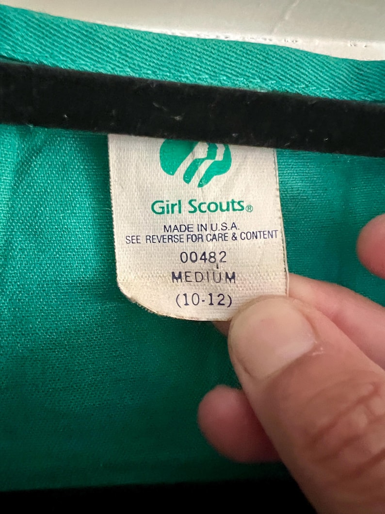 May include: A close-up of a green garment with a tag. The tag reads "Girl Scouts&reg; MADE IN U.S.A." and indicates a medium size (10-12). The garment is a solid green color.