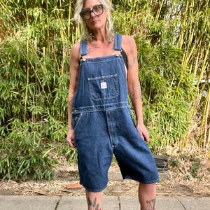 Könnte beinhalten: Dunkelblaue Jeans-Latzshorts mit verstellbaren Trägern und einer Vordertasche. Die Shorts haben eine lockere Passform und werden mit bunten Socken und Sandalen getragen. Im Hintergrund Bambusstängel.