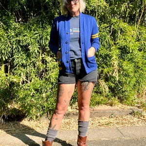 May include: A woman wearing a blue cardigan with yellow stripes on the sleeves, a dark grey t-shirt with the text "Camp 1978" on it, black denim shorts, brown ankle boots, and grey socks. She has short grey hair and is wearing glasses.