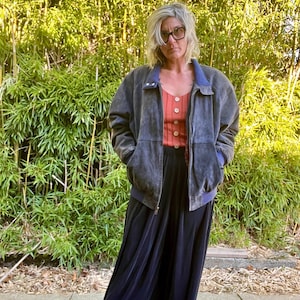 May include: A woman wearing a dark blue suede bomber jacket with a zipper closure and a red button-up shirt underneath. She is wearing black wide-leg pants and has short, gray hair. She is standing in front of a green bamboo background.