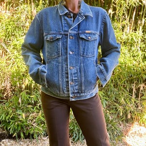May include: A classic blue denim jacket with a button-up front, two chest pockets, and two side pockets. The jacket has a collared neckline and long sleeves. The person is wearing brown pants. The background is a bamboo plant.