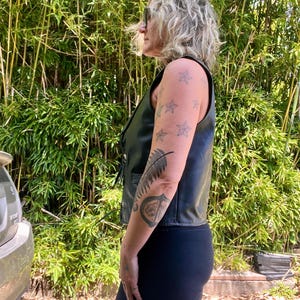 May include: Black leather vest with a sleeveless design. The vest has a button-up front and a V-neck. The person is wearing black leggings. The background is a lush green bamboo forest.
