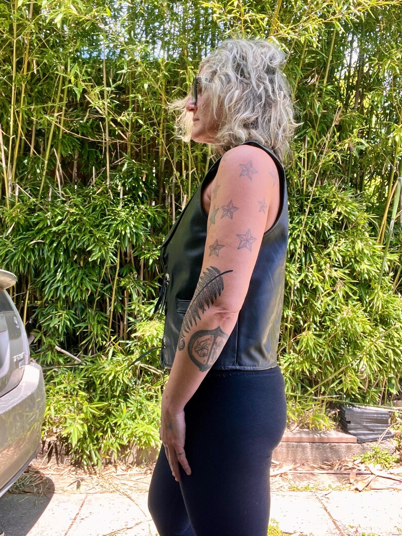 May include: Black leather vest with a sleeveless design. The vest has a button-up front and a V-neck. The person is wearing black leggings. The background is a lush green bamboo forest.
