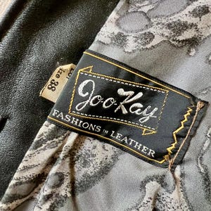 May include: Close-up of a black leather jacket interior. The lining features a gray and white pattern. A black label with gold trim reads "Joo Kay Fashions in Leather". A size 38 tag is visible.
