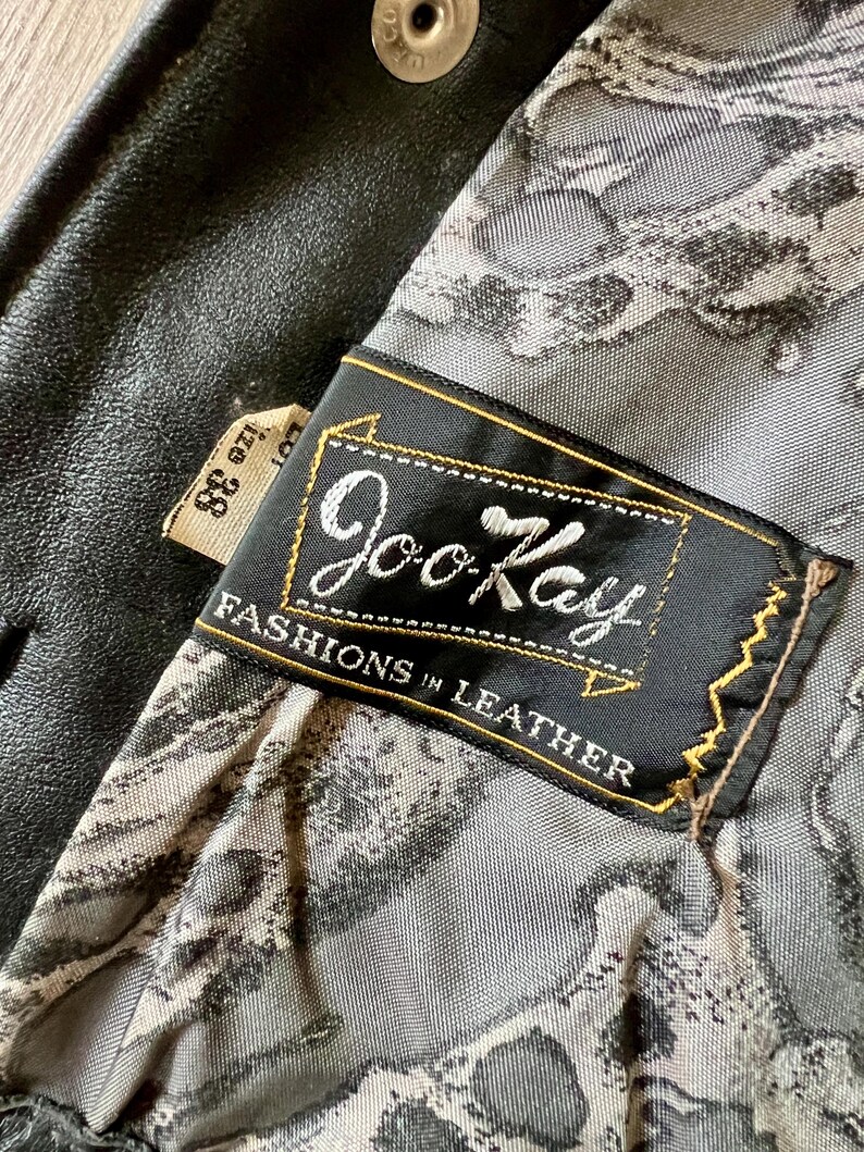 May include: Close-up of a black leather jacket interior. The lining features a gray and white pattern. A black label with gold trim reads "Joo Kay Fashions in Leather". A size 38 tag is visible.