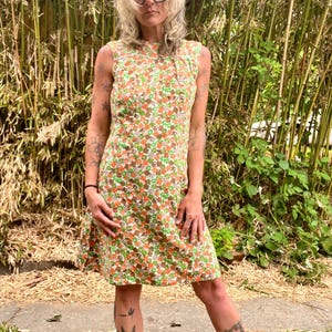 May include: A sleeveless knee-length dress featuring a repeating geometric and floral pattern in shades of orange, green, brown, and white. The dress has a round neckline with white trim detailing and a straight-cut silhouette that falls to the knees.